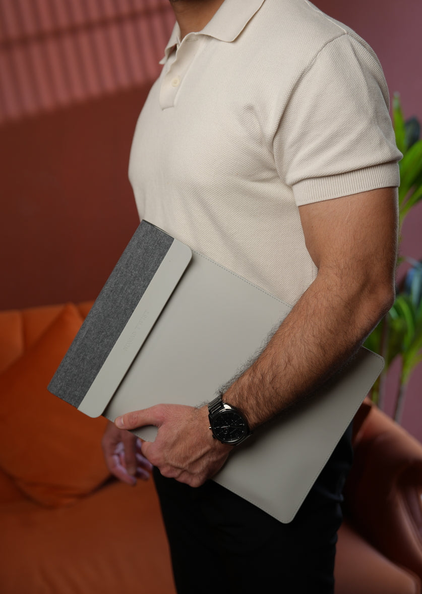 A man in a cream polo shirt holding a light gray laptop sleeve with a textured gray sleeve, standing in a modern room with warm tones.