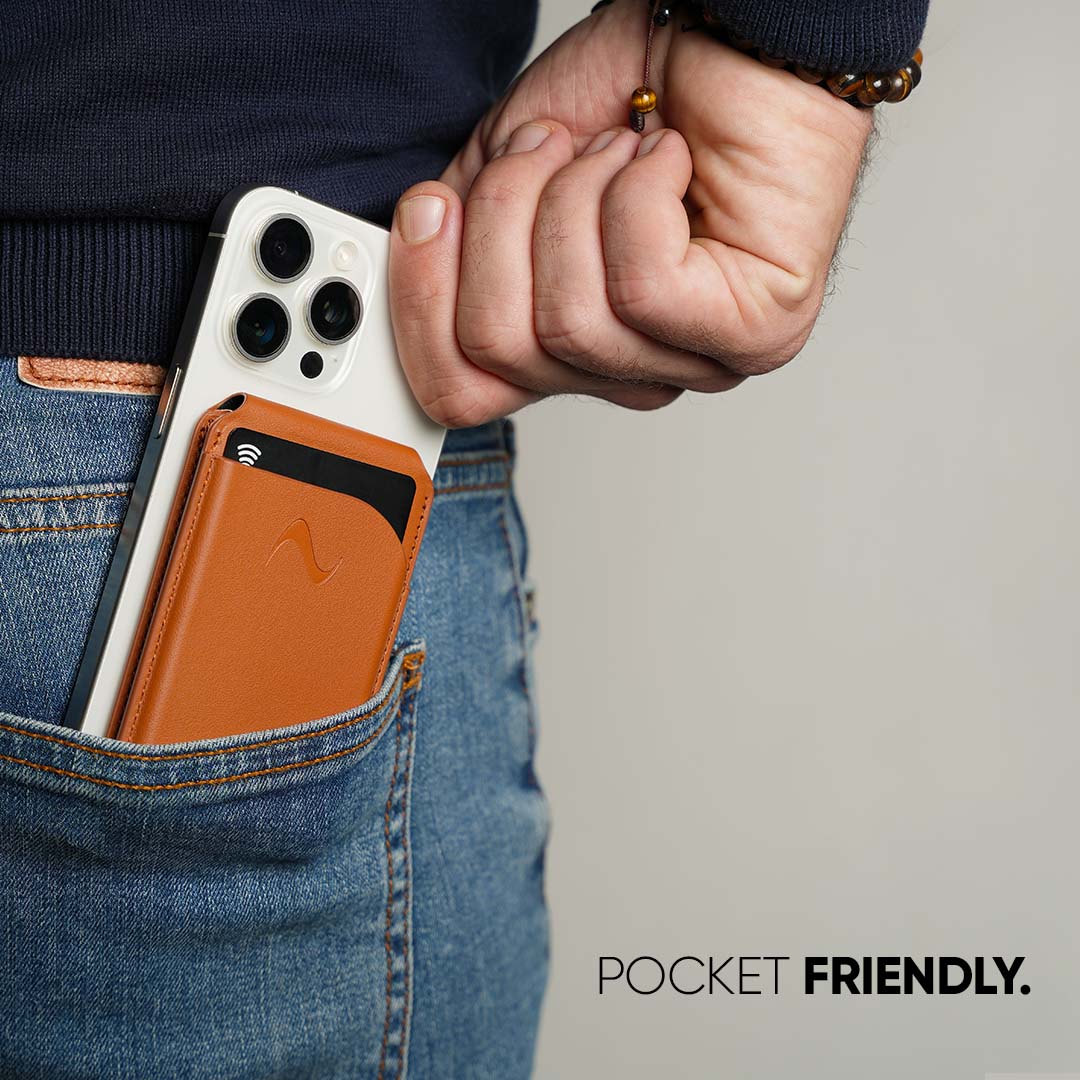 A close-up of a person's hand and torso wearing blue jeans and a dark sweater, with a smartphone tucked into a brown leather card holder wallet in the back pocket. The wallet has a black card slot and a small embossed logo.